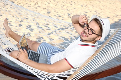 Photo of Young man with laptop resting in hammock at seaside