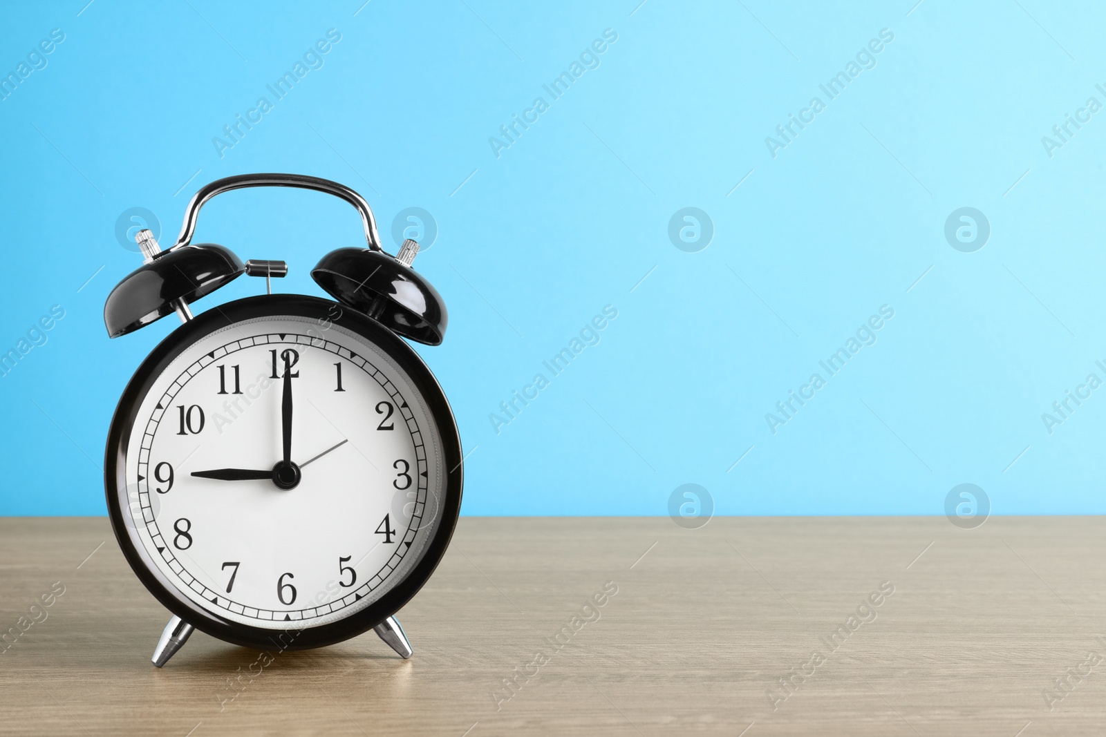 Photo of Black alarm clock on wooden table against light blue background, space for text