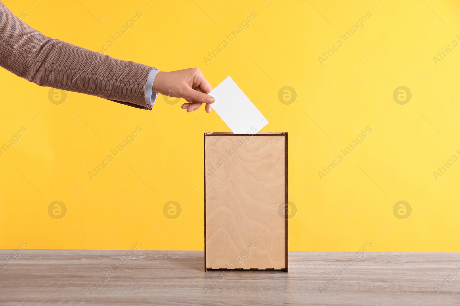 Photo of Woman putting her vote into ballot box on wooden table against orange background, closeup