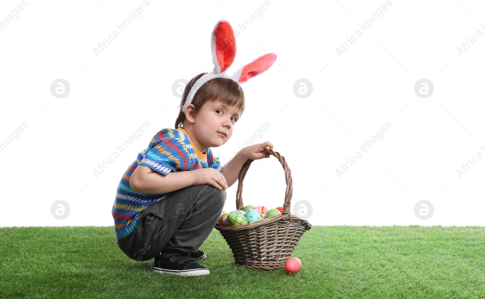 Photo of Cute little boy wearing bunny ears with basket full of dyed Easter eggs on green grass