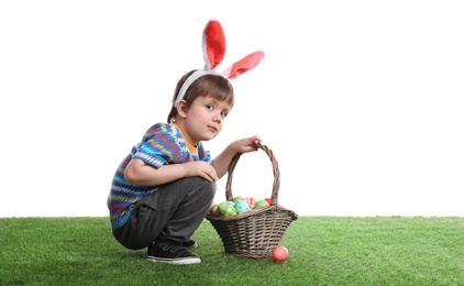 Photo of Cute little boy wearing bunny ears with basket full of dyed Easter eggs on green grass