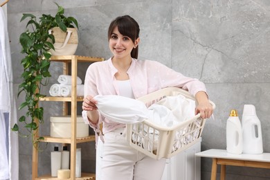 Happy young housewife holding basket with laundry in bathroom