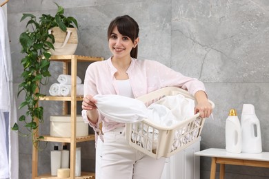 Photo of Happy young housewife holding basket with laundry in bathroom