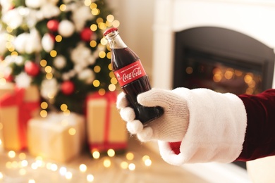 Photo of MYKOLAIV, UKRAINE - JANUARY 18, 2021: Santa Claus holding Coca-Cola bottle in room decorated for Christmas, closeup