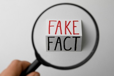 Woman holding magnifying glass over sheet of paper with words Fact and Fake on white background, top view