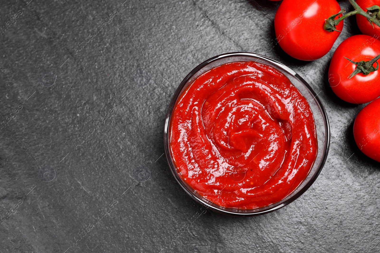 Photo of Organic ketchup in bowl and fresh tomatoes on black table, flat lay with space for text. Tomato sauce