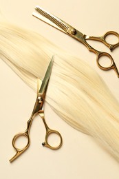 Professional scissors with blonde hair strand on beige background, flat lay