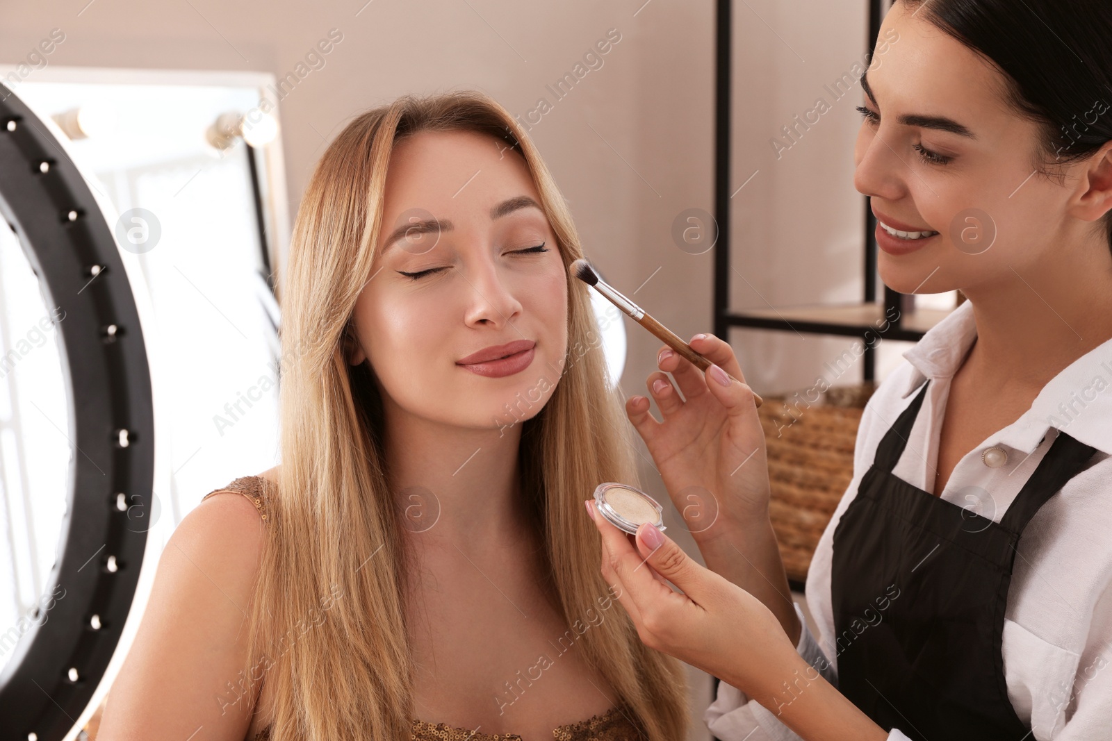 Photo of Professional makeup artist working with beautiful young woman in salon. Using ring lamp