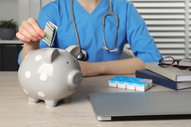 Photo of Doctor putting banknote into piggy bank at wooden table in hospital, closeup. Medical insurance