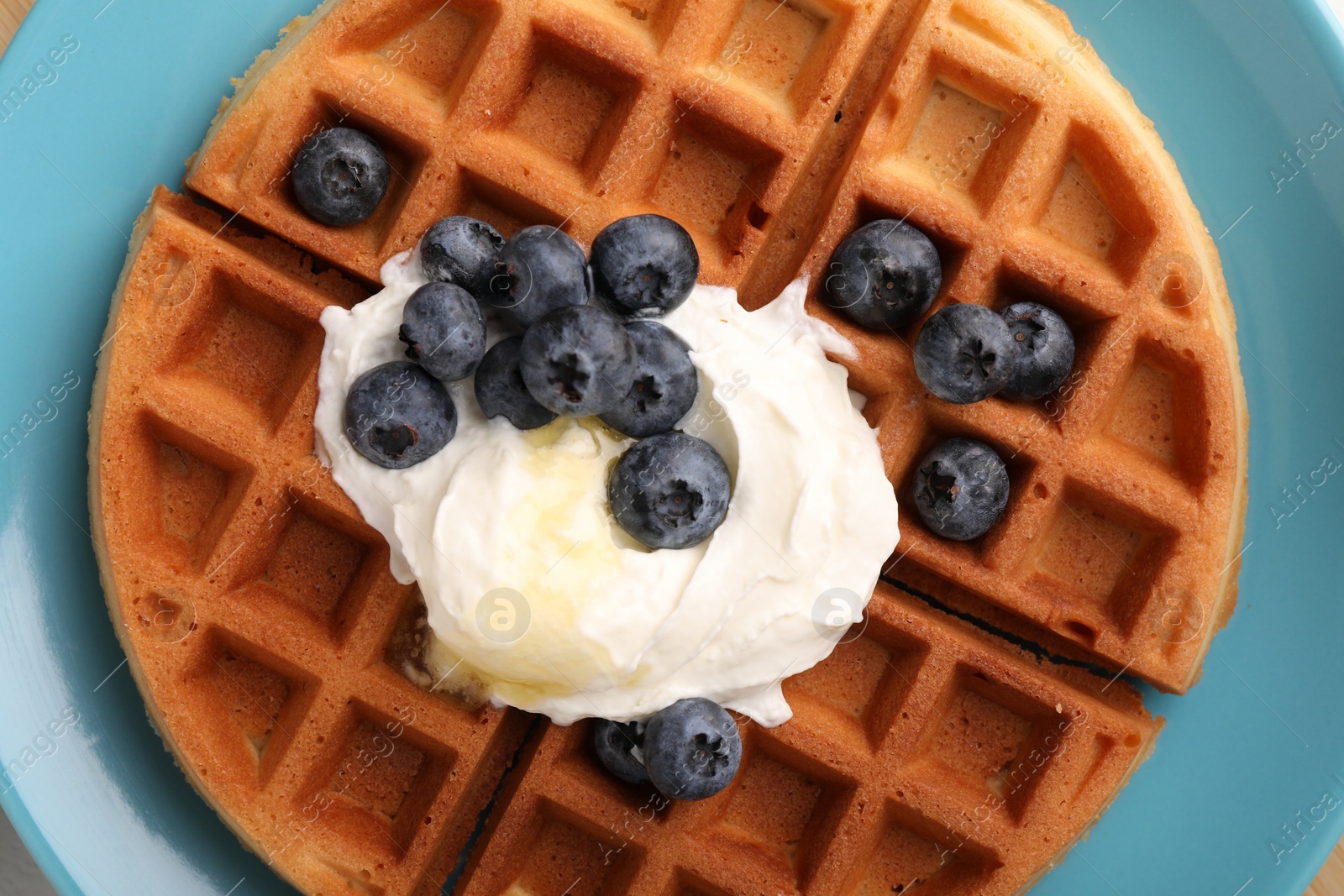Photo of Tasty Belgian waffle with blueberries, honey and whipped cream on plate, top view