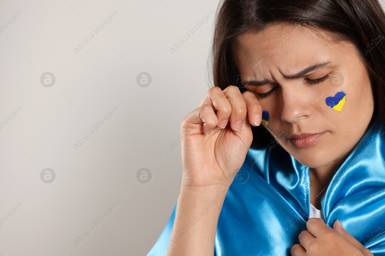 Photo of Sad young woman with face paint and Ukrainian flag on beige background, closeup. Space for text