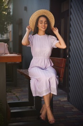Photo of Beautiful young woman in stylish violet dress and straw hat sitting on bench outdoors