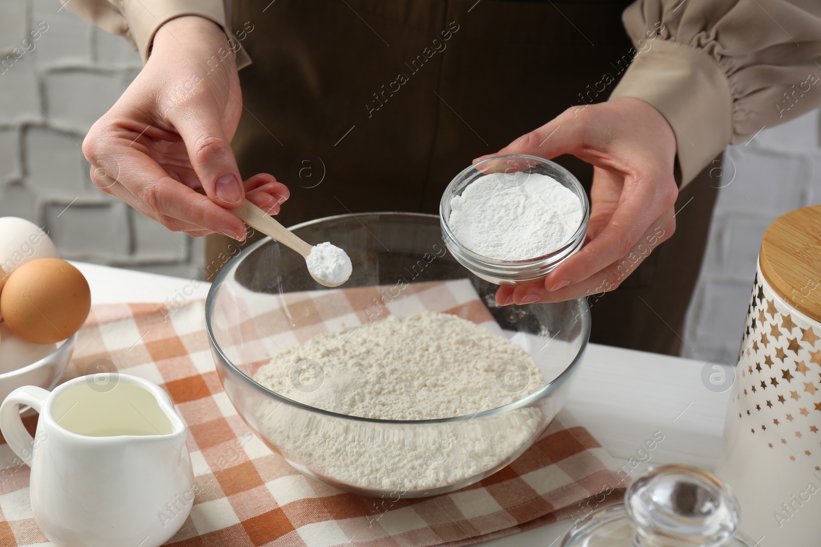 Photo of Woman with spoon and bowl of baking powder at white table, closeup