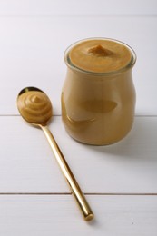 Photo of Spoon and glass jar of tasty mustard sauce on white wooden table, closeup