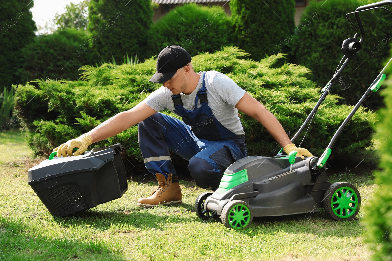 Photo of Cleaning lawn mower. Young man detaching grass catcher from device in garden