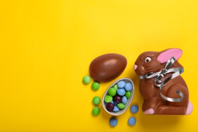 Photo of Chocolate Easter bunny, halves of egg and candies on yellow background, flat lay. Space for text