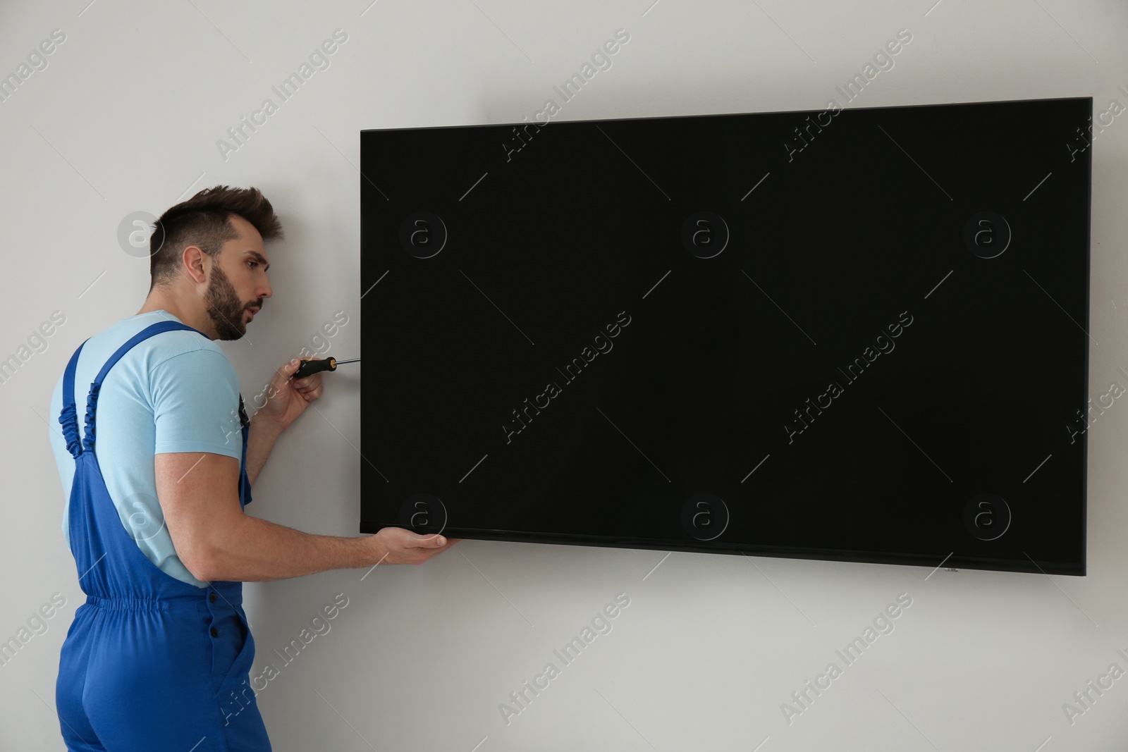 Photo of Professional technician with screwdriver installing modern flat screen TV on wall indoors