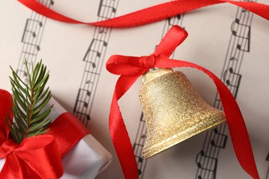 Photo of Golden shiny bell with red bow and gift box on music sheet, flat lay. Christmas decoration