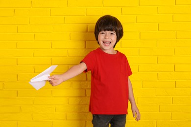 Photo of Cute little boy with paper plane near yellow brick wall