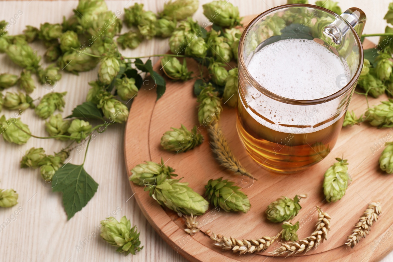 Photo of Mug with beer, fresh hops and ears of wheat on light wooden table