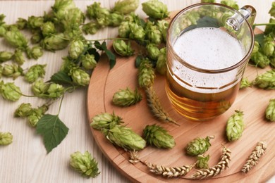 Photo of Mug with beer, fresh hops and ears of wheat on light wooden table