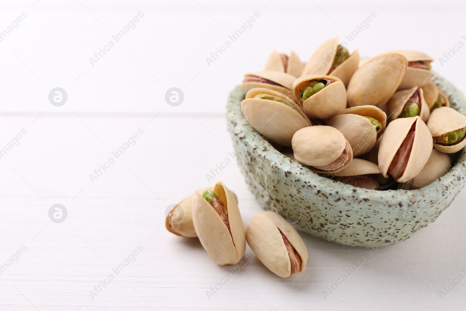 Photo of Delicious pistachios in bowl on white table, closeup. Space for text