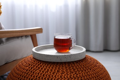 Tray with cup of tea on stylish knitted pouf in room. Interior design