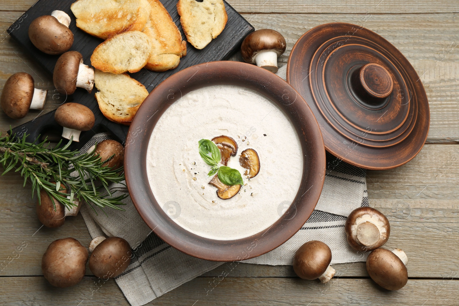 Photo of Tasty homemade mushroom soup in ceramic pot, croutons, fresh champignons and rosemary on wooden table, flat lay