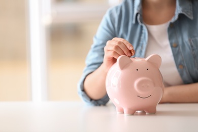 Photo of Woman putting coin into piggy bank at table indoors, closeup. Space for text
