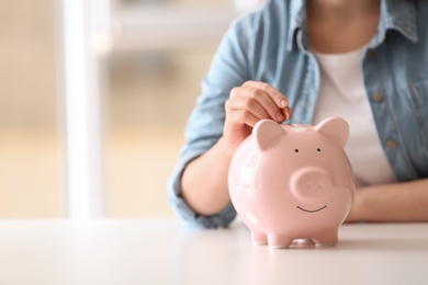 Woman putting coin into piggy bank at table indoors, closeup. Space for text