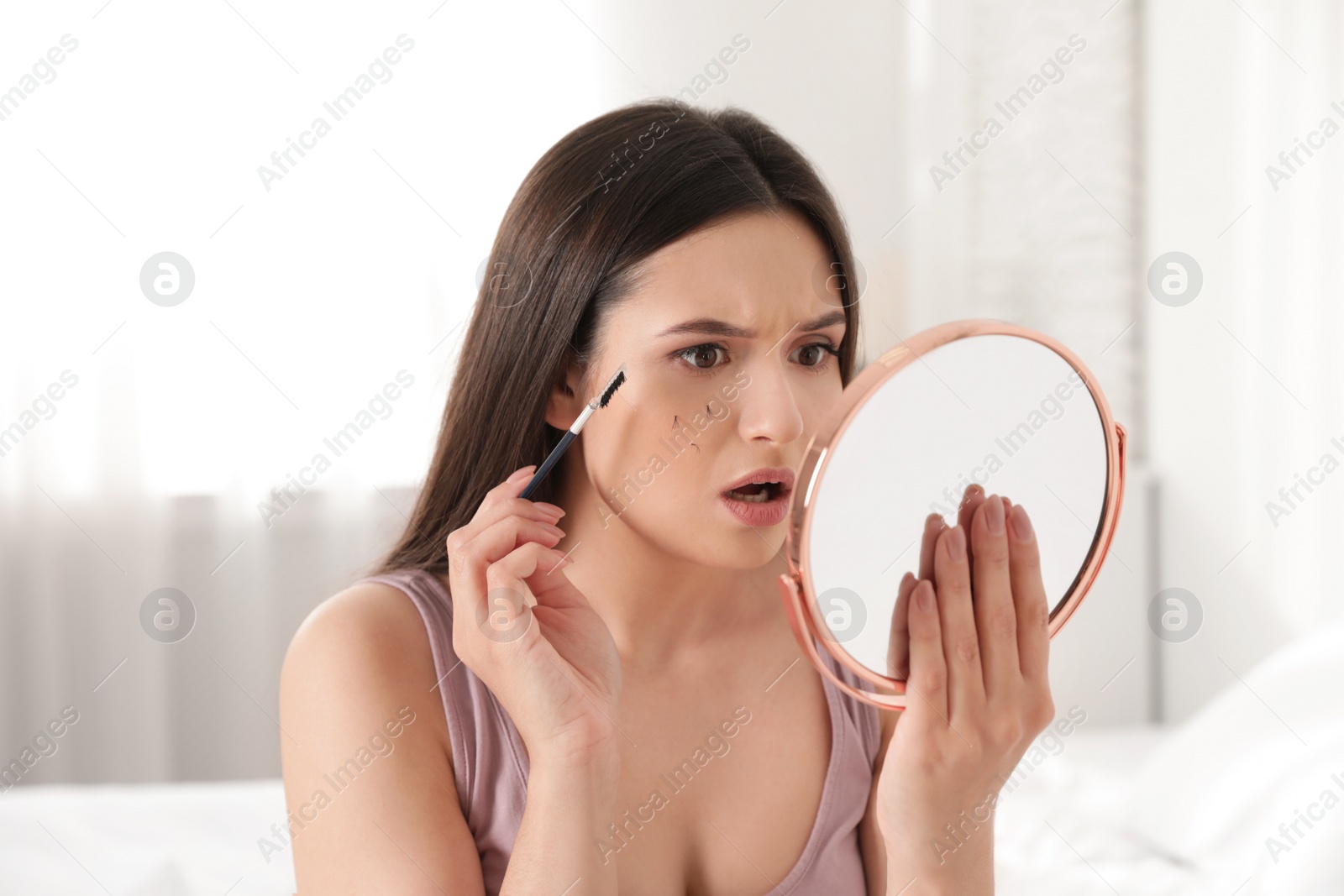 Photo of Beautiful woman with fallen eyelashes and cosmetic brush looking into mirror indoors