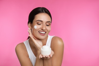 Young woman applying facial cream on pink background