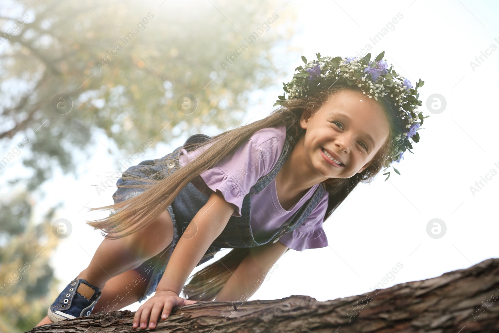 Photo of Cute little girl wearing flower wreath on tree outdoors. Child spending time in nature