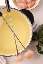 Fondue pot with oil, forks, raw meat pieces and parsley on white wooden table, flat lay