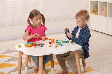 Little children playing with wooden pieces and string for threading activity at white table indoors. Developmental toys