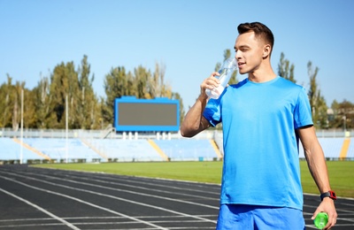 Young sporty man drinking water from bottle at stadium on sunny day. Space for text