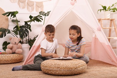 Photo of Cute little children reading book near toy wigwam at home