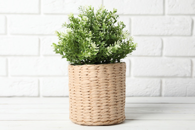 Photo of Beautiful artificial plant in wicker flower pot on white wooden table near brick wall