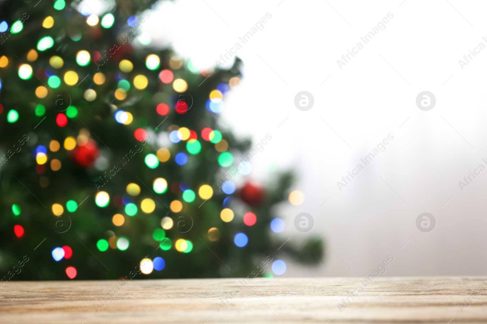 Photo of Blurred view of fir tree with glowing Christmas lights near table indoors. Festive mood