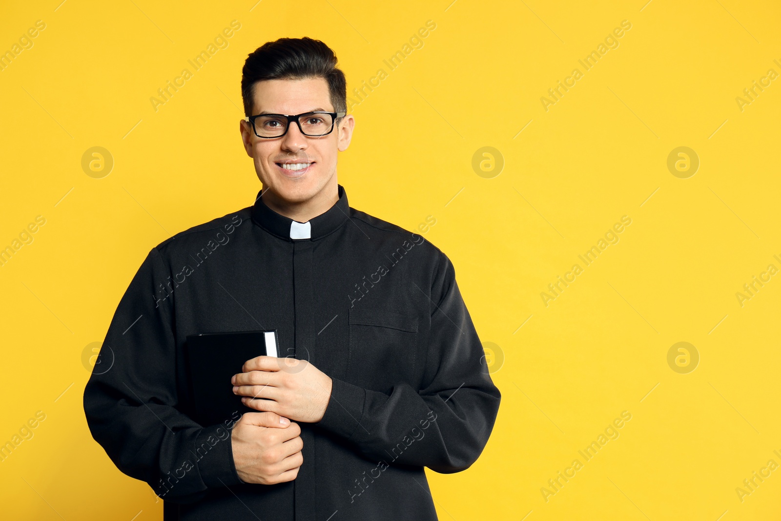 Photo of Priest in cassock with Bible on yellow background. Space for text