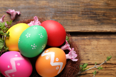 Colorful Easter eggs in decorative nest with flowers on wooden background, closeup