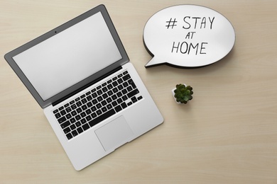 Laptop, houseplant and speech bubble with hashtag STAY AT HOME on wooden background, flat lay. Message to promote self-isolation during COVID‑19 pandemic