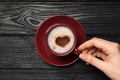 Photo of Woman holding cup of aromatic coffee with heart shaped decoration at black wooden table, top view