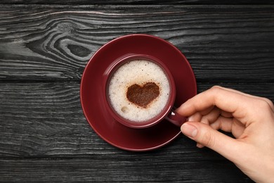 Woman holding cup of aromatic coffee with heart shaped decoration at black wooden table, top view