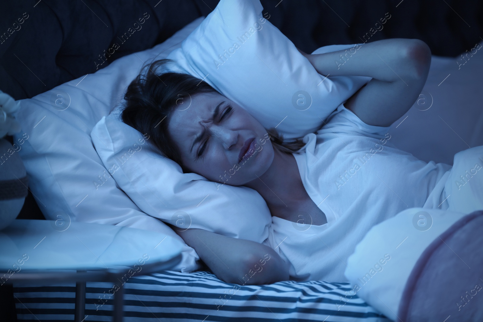 Photo of Young woman covering ears with pillow while trying to sleep in bed at night