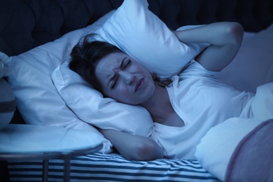 Photo of Young woman covering ears with pillow while trying to sleep in bed at night