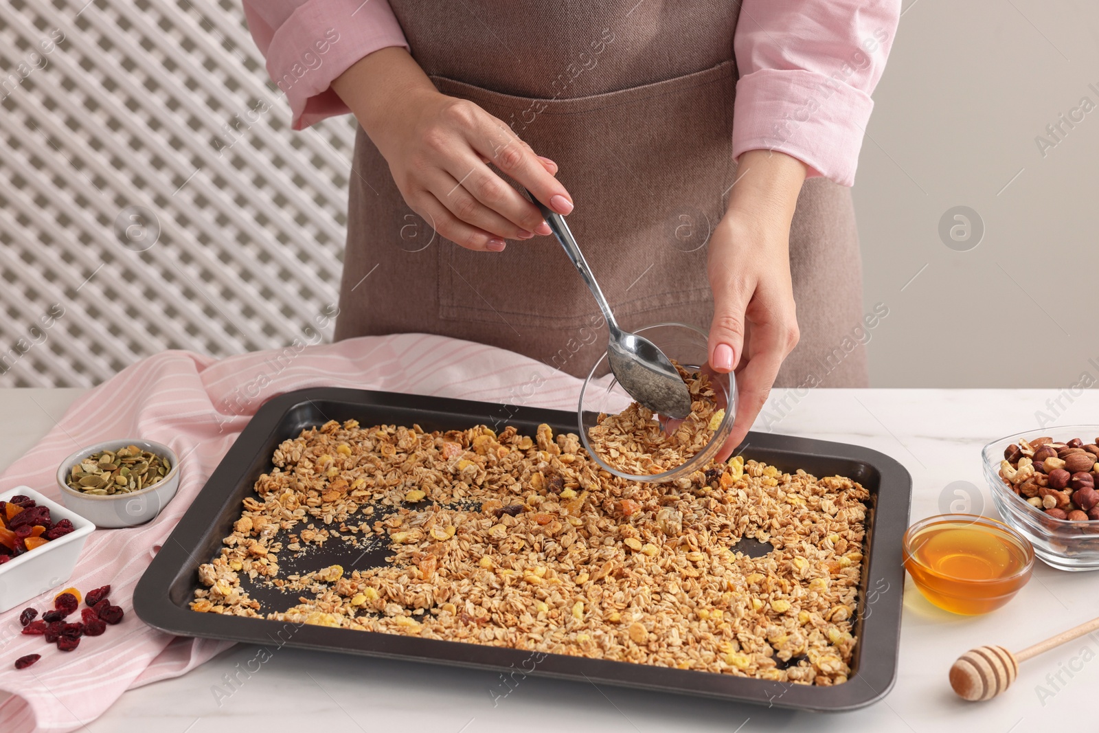 Photo of Woman putting granola from baking tray into glass bowl at white table, closeup