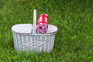 Photo of Rolled checkered tablecloth with picnic basket on green grass outdoors, space for text