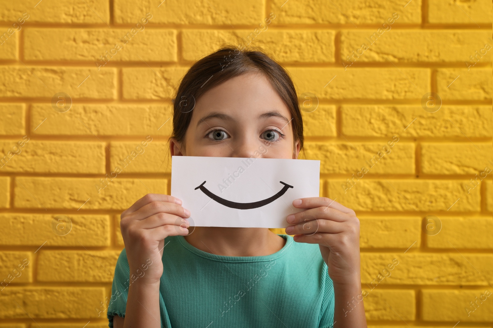 Photo of Little girl holding sheet of paper with smile against yellow brick wall