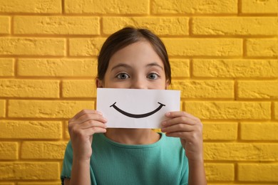 Little girl holding sheet of paper with smile against yellow brick wall